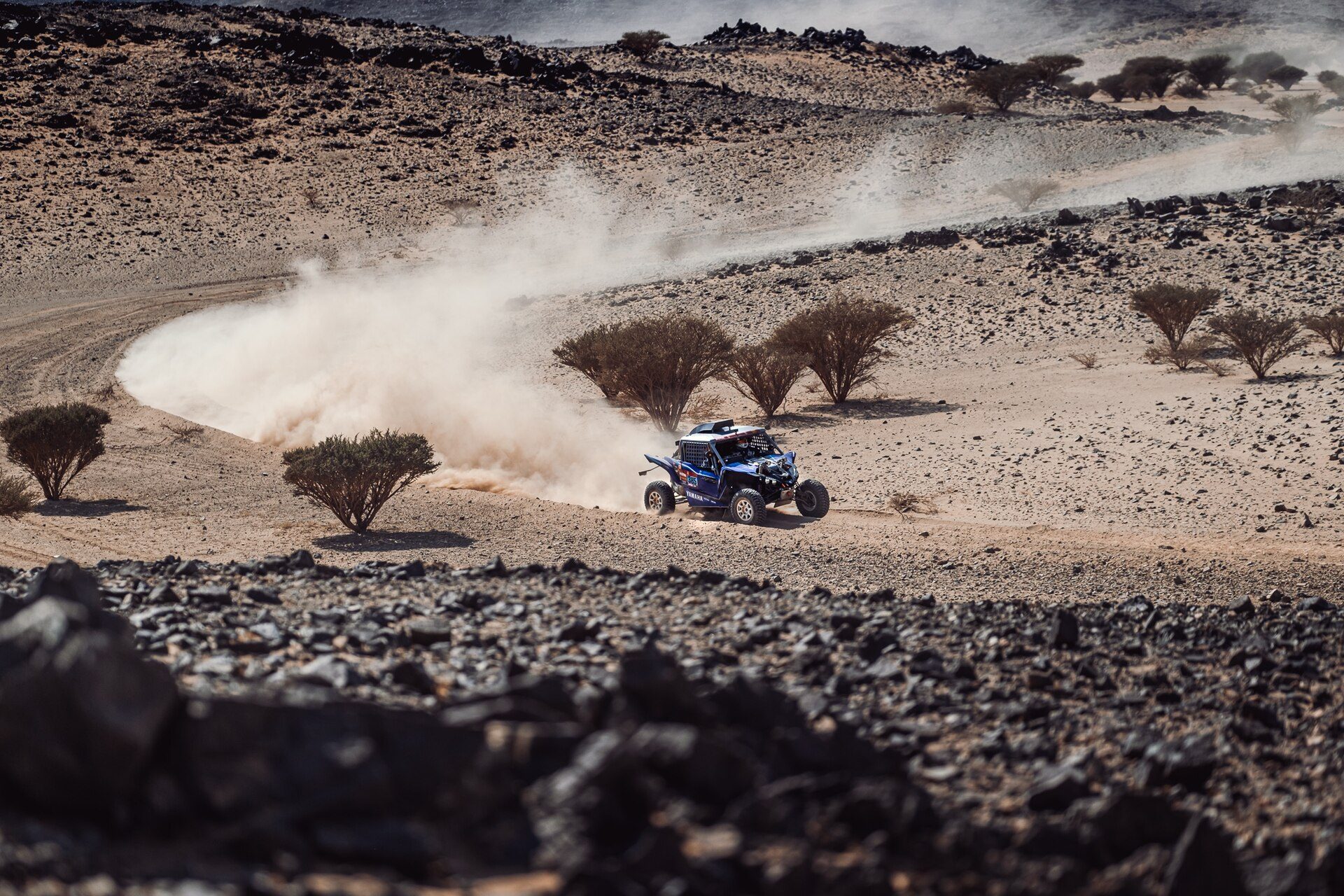 Rally vehicle crossing a stony desert plateau in a cloud of dust